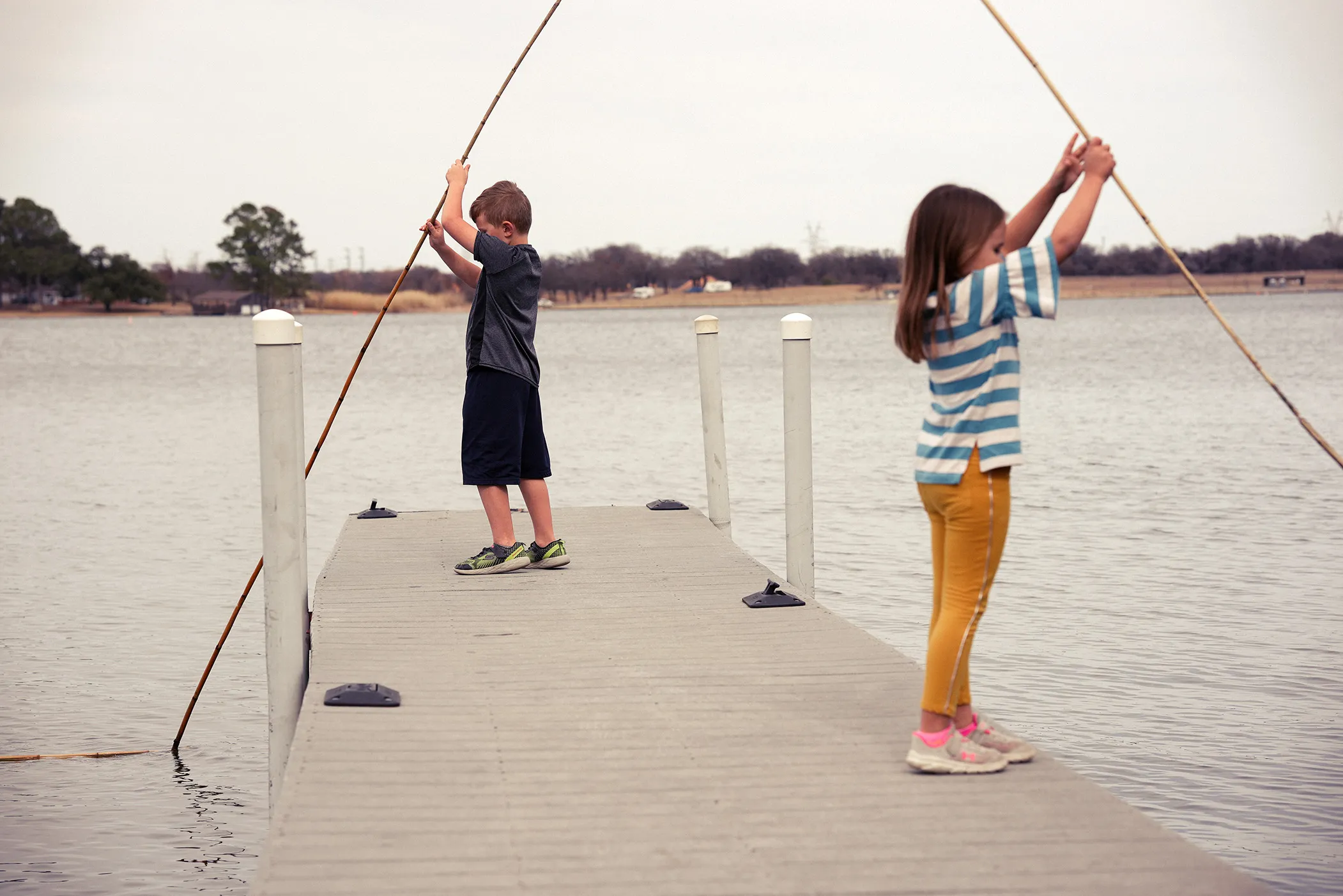 Two children holding bamboo poles at the end of a lake dock