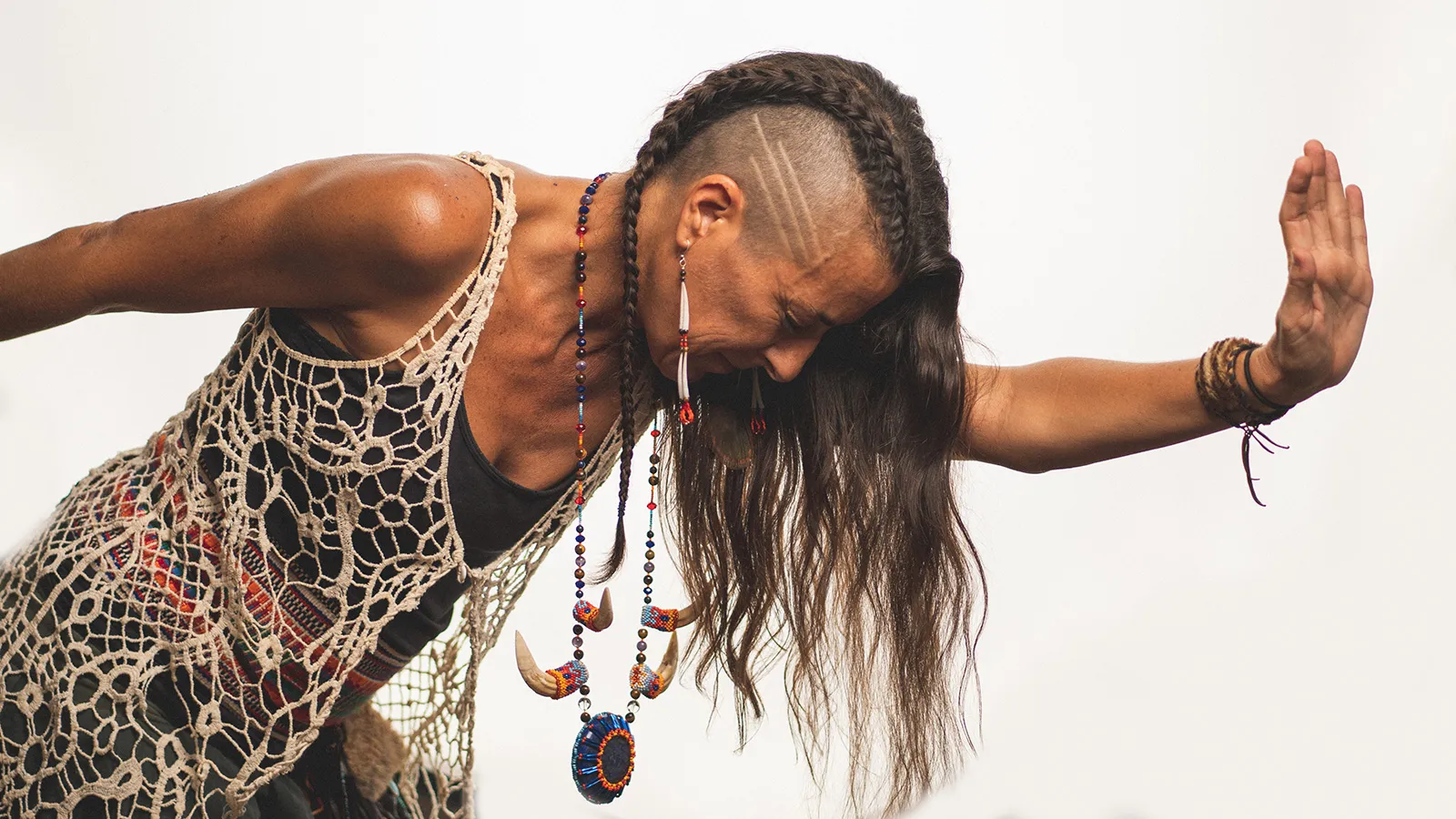 A dancer bowed forward, braids and beadwork against a white ground